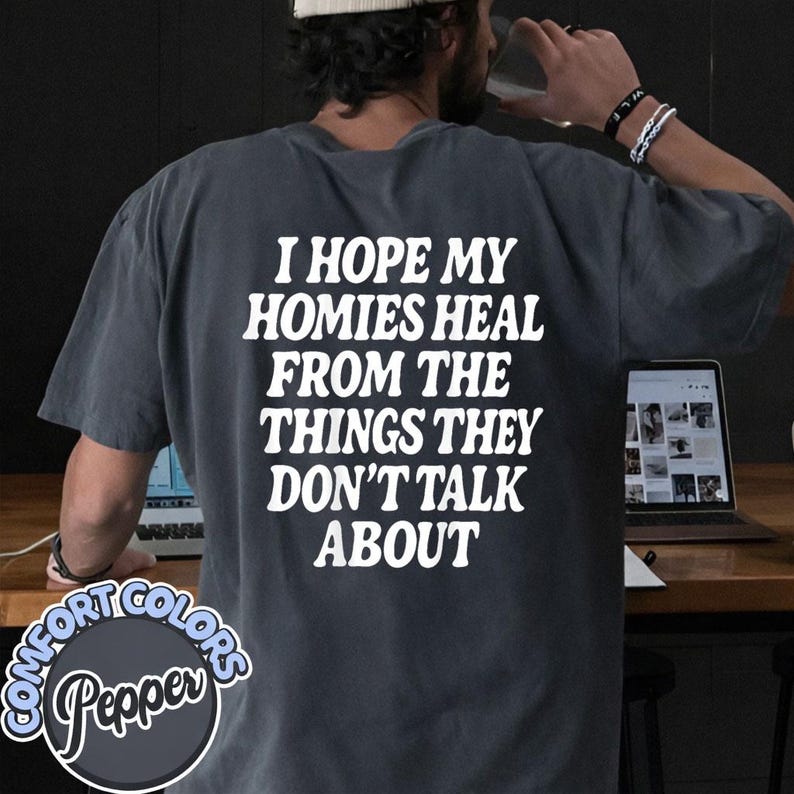 May include: A charcoal gray short-sleeve t-shirt featuring white text on the back that reads, I HOPE MY HOMIES HEAL FROM THE THINGS THEY DON'T TALK ABOUT. The shirt has a relaxed fit and a solid color design with a circular logo in the corner.