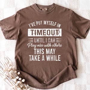 May include: A brown t-shirt with the text "I'VE PUT MYSELF IN TIMEOUT UNTIL I CAN Play nice with others THIS MAY TAKE A WHILE" in white. The shirt has a crew neck and short sleeves. The fabric appears soft and slightly faded.