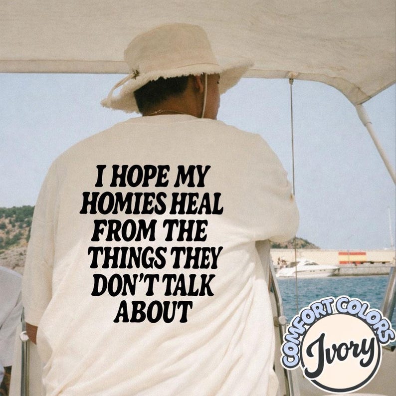 May include: An ivory-colored t-shirt featuring black text on the back that reads, I HOPE MY HOMIES HEAL FROM THE THINGS THEY DON'T TALK ABOUT. The shirt is worn with a light-colored, wide-brimmed hat with a frayed edge and a chin strap.