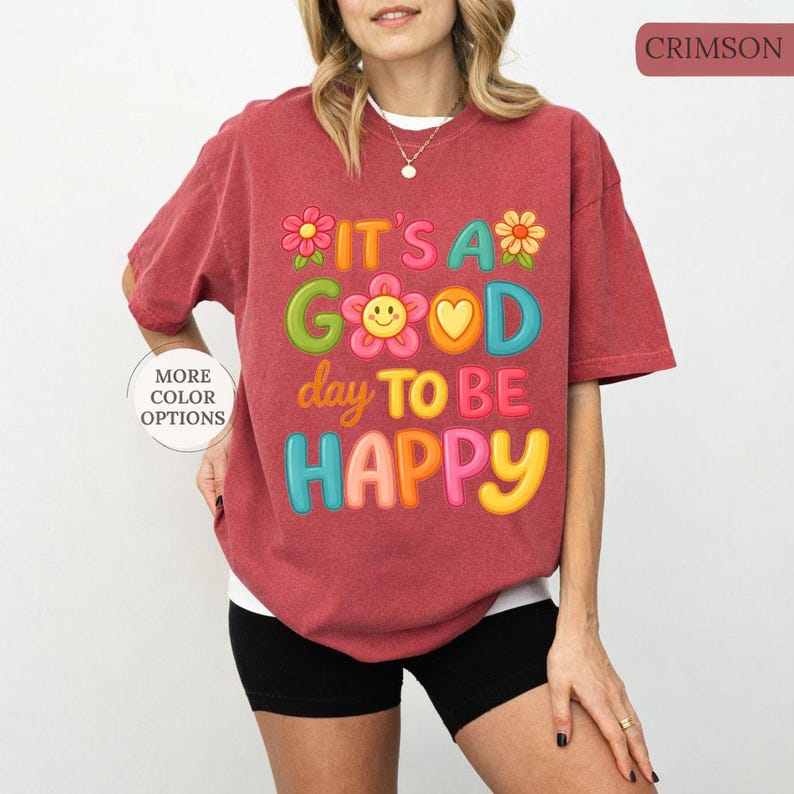 May include: Crimson-colored t-shirt with the text "IT'S A GOOD day TO BE HAPPY" in colorful, playful letters and flower graphics. The shirt is a relaxed fit and has short sleeves. The image also shows a button that says "MORE COLOR OPTIONS".