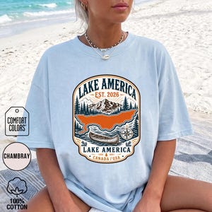 May include: A light blue t-shirt featuring a graphic print with the text LAKE AMERICA EST. 2026. The design includes a mountain range, pine trees, a canoe on water, a compass rose, and the text LAKE AMERICA CANADA / USA in a rounded frame.