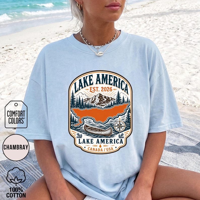 May include: A light blue t-shirt featuring a graphic print with the text LAKE AMERICA EST. 2026. The design includes a mountain range, pine trees, a canoe on water, a compass rose, and the text LAKE AMERICA CANADA / USA in a rounded frame.