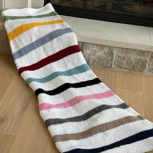 May include: A white knitted blanket with horizontal stripes in various colours, including yellow, blue, red, and black. The blanket is lying on a wooden floor.