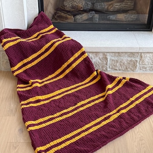 Può includere: Una coperta a maglia a righe bordeaux e oro. La coperta presenta spesse strisce orizzontali dorate su uno sfondo bordeaux. La coperta è piegata, mostrando la sua consistenza e il suo design.
