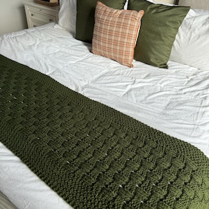 May include: A dark green knitted bed runner with a textured pattern, placed on a white bed. The bed is adorned with green and beige pillows, creating a cozy aesthetic. The runner is approximately 183 cm long.