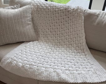 May include: A white knitted blanket with a textured pattern is draped over a beige sofa. A matching pillow rests on the sofa. The blanket's design features a repeating pattern, and the edges are finished with a ribbed border.