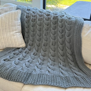 Puede incluir: Una manta gris tejida con un diseño de punto de cable. La manta está sobre un sofá, mostrando su textura y patrón. Los bordes tienen un borde acanalado. Una almohada es visible en el lado izquierdo de la imagen.
