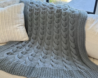May include: A gray knitted blanket with a cable knit design. The blanket is draped over a sofa, showcasing its texture and pattern. The edges have a ribbed border. A pillow is visible on the left side of the image.