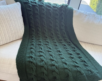 May include: A dark green knitted blanket with a cable knit design is draped over a light-colored sofa. The blanket has a thick, textured appearance with a knitted border. The sofa has two light-colored pillows.