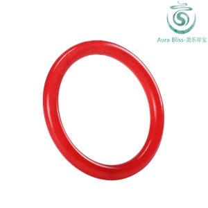 May include: A bright red, circular bangle bracelet. The smooth, polished surface reflects light, highlighting the rich colour. The Aura Bliss logo is visible in the upper right corner.