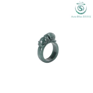 May include: A dark green jade ring featuring a carved lion design. The ring is a solid band with the lion figure resting on top. The ring is set against a white background.
