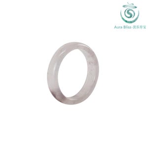 Puede incluir: Un anillo de piedra preciosa rosa pálido translúcido. El anillo es una banda sólida con una superficie lisa y pulida. El logotipo de Aura Bliss está en la esquina superior derecha.