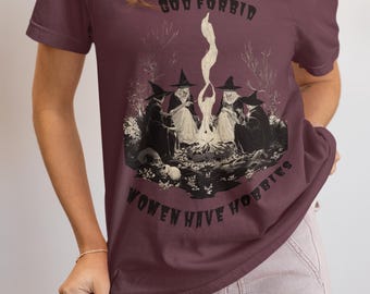 Puede incluir: Una camiseta de manga corta color granate oscuro con una ilustración gráfica de cuatro figuras con sombreros puntiagudos sentadas alrededor de una fogata. El texto "GOD FORBID" aparece arriba y "WOMEN HAVE HOBBIES" aparece debajo.