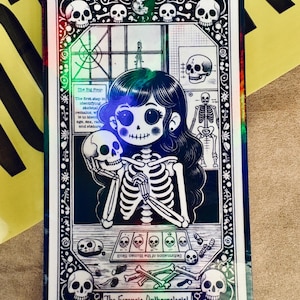 Puede incluir: Una tarjeta holográfica con una ilustración de dibujos animados de una niña con un disfraz de esqueleto sosteniendo una calavera. La tarjeta tiene un borde en blanco y negro con motivos de calaveras y el texto "The Forensic Anthropologist" y "12. The Forensic Anthropologist".