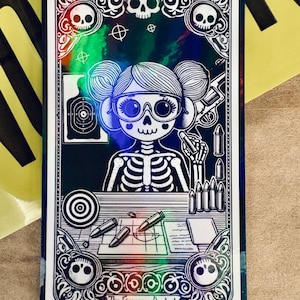 May include: A tarot card illustration featuring a skeleton figure with glasses, a gun, and bullets. The card has a decorative border with skulls and the text "The Firearms Analyst". The card is labeled "8. The Firearms Analyst - Female".