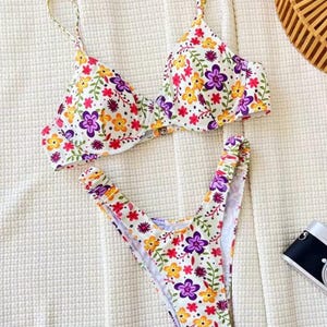 May include: A two-piece bikini set with a white background and a vibrant floral pattern. The bikini top has adjustable straps and a clasp closure. The floral design includes purple, yellow, and red flowers with green leaves.