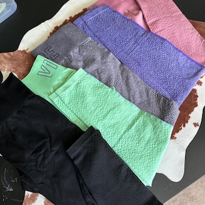 May include: An assortment of workout leggings in various colors, including black, mint green, gray, purple, and pink. The leggings have a textured pattern and the word "Vibe" is visible on some of them. The leggings are arranged on a patterned surface.
