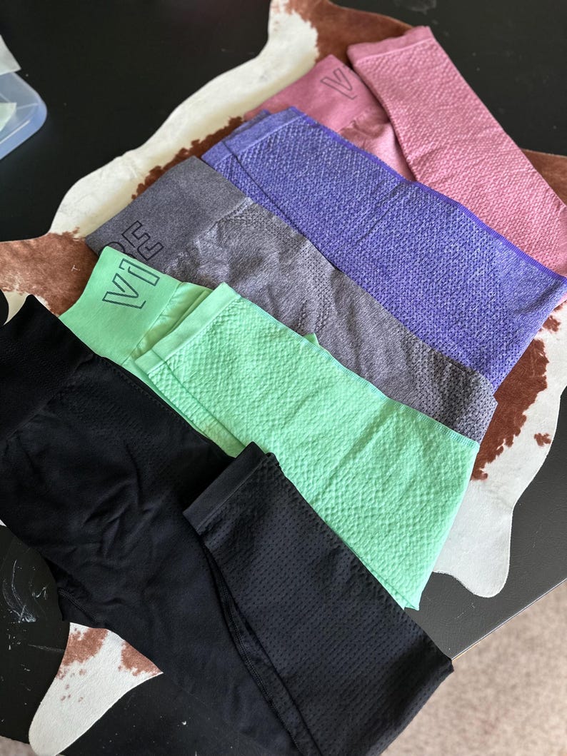 May include: An assortment of workout leggings in various colors, including black, mint green, gray, purple, and pink. The leggings have a textured pattern and the word "Vibe" is visible on some of them. The leggings are arranged on a patterned surface.