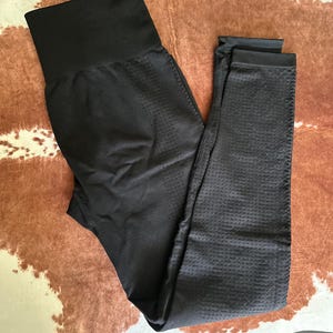 May include: Black leggings with a high waistband. The leggings have a textured, perforated pattern on the legs and a smooth, solid waistband. The leggings are made of a stretchy material and are designed for athletic activities.