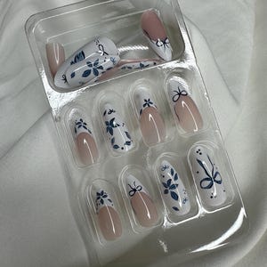 May include: A set of artificial nails in a clear plastic container. The nails feature a variety of designs, including a nude base with a white tip and a black bow, and white nails with blue floral patterns. The nails are oval-shaped.