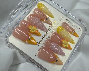 May include: A set of handmade, almond-shaped press-on nails in a clear plastic case. The nails feature a nude base with yellow tips, floral accents, and small silver embellishments. Text on the case reads "Handmade Nails".