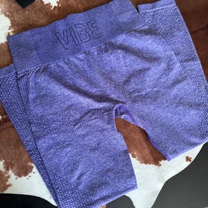May include: Purple workout leggings with the word "VIBE" across the waistband. The leggings are a solid color with a textured knit design. The leggings are folded and laying on a patterned surface.