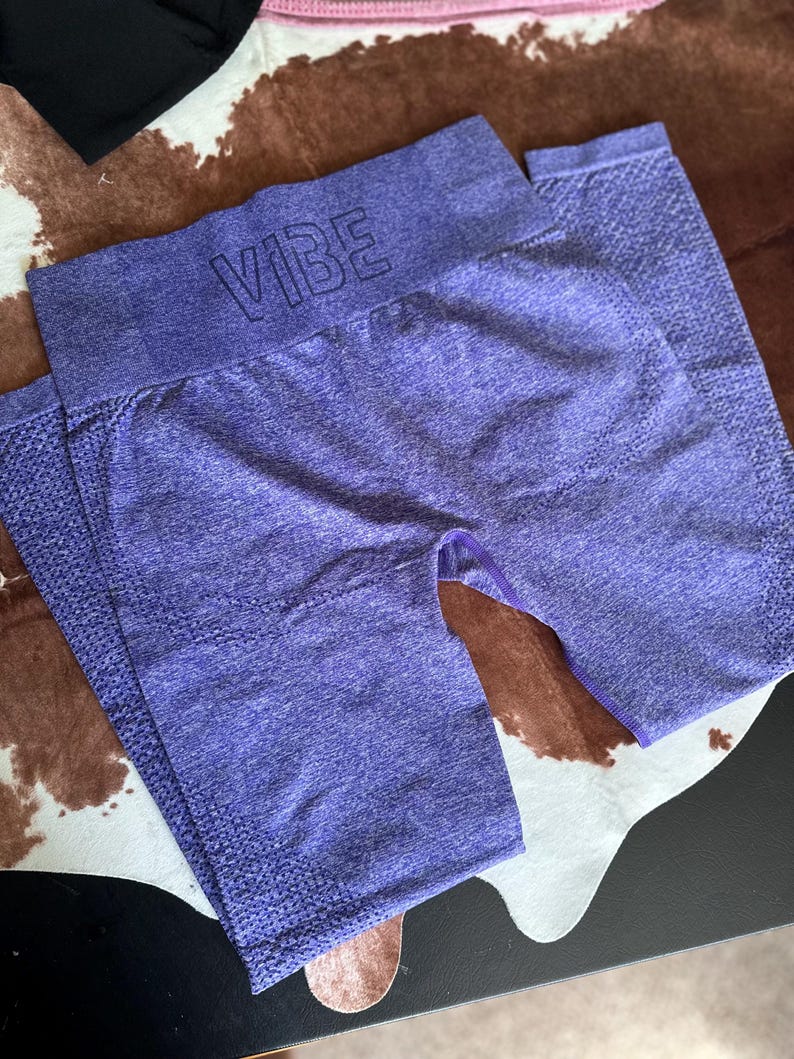 May include: Purple workout leggings with the word "VIBE" across the waistband. The leggings are a solid color with a textured knit design. The leggings are folded and laying on a patterned surface.