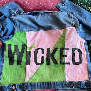 May include: A blue denim jacket with a vibrant back design. The design includes the word "WICKED" in black, set against green and pink geometric shapes. A green witch silhouette is also visible.