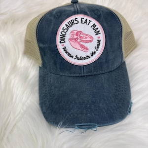 May include: A distressed navy blue trucker hat with a tan mesh back. The front features a circular patch with the text "DINOSAURS EAT MAN" and "Woman Inherits the Earth" surrounding a pink dinosaur skull.