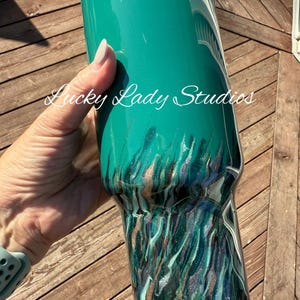 K&ouml;nnte beinhalten: Ein t&uuml;rkisfarbener Becher mit einem metallischen, flie&szlig;enden Design in T&uuml;rkis-, Silber- und Braunt&ouml;nen. Der Text "Lucky Lady Studios" ist kursiv auf die Seite des Bechers geschrieben. Der Becher wird vor einem h&ouml;lzernen Hintergrund gehalten.