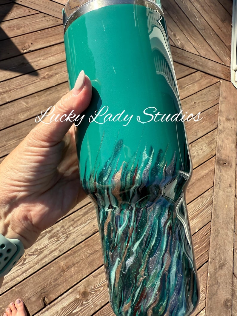 K&ouml;nnte beinhalten: Ein t&uuml;rkisfarbener Becher mit einem metallischen, flie&szlig;enden Design in T&uuml;rkis-, Silber- und Braunt&ouml;nen. Der Text "Lucky Lady Studios" ist kursiv auf die Seite des Bechers geschrieben. Der Becher wird vor einem h&ouml;lzernen Hintergrund gehalten.