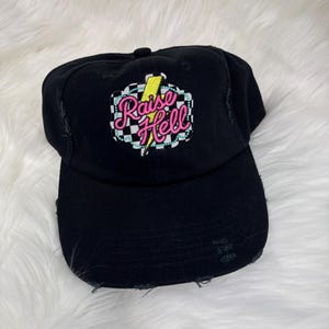 May include: Black baseball cap with distressed edges. The cap features an embroidered design with the words "Raise Hell" in pink and yellow, set against a checkered background with a yellow lightning bolt.