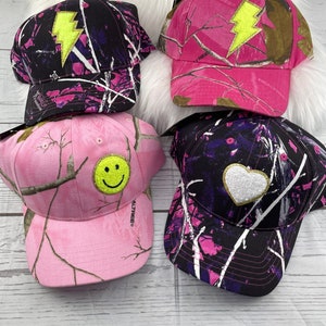 May include: Four pink and black camouflage baseball caps with different embroidered patches. The patches include a lightning bolt, a smiley face, and a heart.