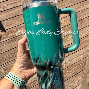 K&ouml;nnte beinhalten: Ein tealfarbener Stanley-Becher mit Griff und transparentem Deckel. Der Becher hat ein Gras- und Wassermotiv in Teal-, Schwarz- und Silbert&ouml;nen. Der Text "Lucky Lady Studios" ist auf dem Becher geschrieben.