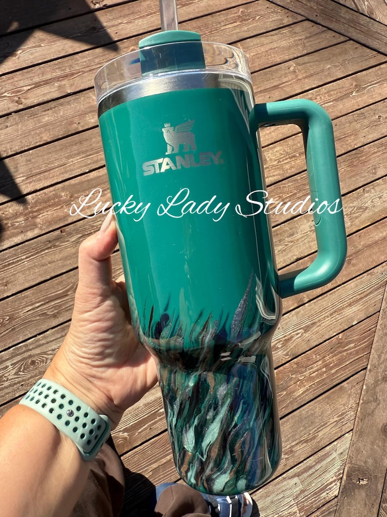 K&ouml;nnte beinhalten: Ein tealfarbener Stanley-Becher mit Griff und transparentem Deckel. Der Becher hat ein Gras- und Wassermotiv in Teal-, Schwarz- und Silbert&ouml;nen. Der Text "Lucky Lady Studios" ist auf dem Becher geschrieben.