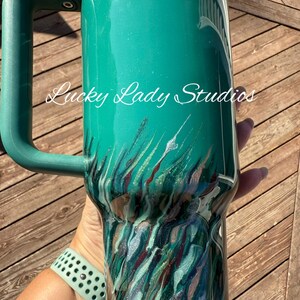 K&ouml;nnte beinhalten: Ein tealfarbener Becher mit Griff, mit einem marmorierten Design in den Farbt&ouml;nen Teal, Silber und Burgunder. Der Text "Lucky Lady Studios" ist in wei&szlig;er Schrift auf der Seite des Bechers geschrieben. Eine tealfarbene Armbanduhr ist sichtbar.