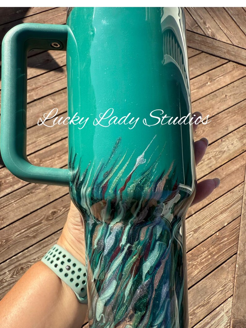 K&ouml;nnte beinhalten: Ein tealfarbener Becher mit Griff, mit einem marmorierten Design in den Farbt&ouml;nen Teal, Silber und Burgunder. Der Text "Lucky Lady Studios" ist in wei&szlig;er Schrift auf der Seite des Bechers geschrieben. Eine tealfarbene Armbanduhr ist sichtbar.