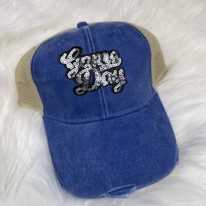 May include: A blue distressed baseball cap with a tan mesh back. The front of the cap features the sequined text "Lazy Day" in a cursive font. The cap has a curved brim and a button on top.