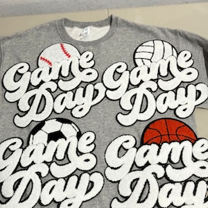 May include: A gray sweatshirt featuring four sports balls: baseball, volleyball, soccer ball, and basketball. Each ball is paired with the words "Game Day" in a white, fluffy, cursive font, creating a sporty theme.