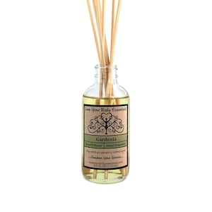 May include: A clear glass bottle with a brown label that reads "Love Your Body Essentials Gardenia Reed Diffuser | Home Fragrance Flip reeds as needed to refresh scent...Awaken Your Senses...loveyourbodyessentials.com 4.0 FL OZ (118 ml)". The bottle is filled with a yellow liquid and has wooden sticks inserted into it.