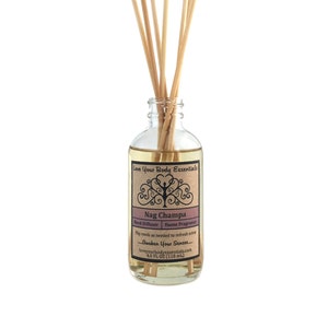 May include: A clear glass bottle with a brown label that reads "Love Your Body Essentials Nag Champa Reed Diffuser Home Fragrance Flip reeds as needed to refresh scent... Awaken Your Senses... loveyourbodyessentials.com 4.0 FL OZ (118 ml)". The bottle is filled with a light yellow liquid and has wooden sticks inserted into it.