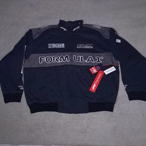 Può includere: Giacca Formula 1 blu navy e grigia con cerniera intera, con la scritta "FORMULA 1" sul petto. La giacca presenta la scritta "FAST IS NOT ENOUGH" e "INTL" sul davanti, con un'etichetta Formula 1 allegata.