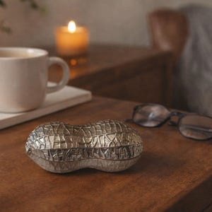 May include: A silver-coloured peanut-shaped decorative object sits on a wooden surface. A white mug, a candle, and a pair of glasses are also on the table. The peanut-shaped object has a textured surface.