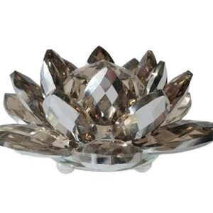 May include: A decorative lotus flower sculpture made of faceted, smoky-colored crystals. The crystal petals are arranged in a circular pattern, with a central, diamond-shaped crystal. The sculpture sits on a clear, round base.