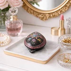 May include: A black, heart-shaped trinket box with a floral design sits on a marble tray with a gold trim. The box is surrounded by beauty products and jewellery, including a perfume bottle, lipstick, and a pearl necklace.