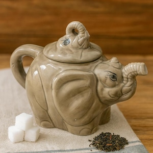 Puede incluir: Tetera de cerámica gris claro con forma de elefante. La tetera tiene un pico en forma de trompa, un asa y una tapa con un adorno de cabeza de elefante. Junto a la tetera hay tres cubos de azúcar y un montón de hojas de té sueltas. La tetera está sobre un paño a rayas.