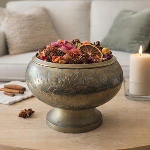 Könnte beinhalten: Eine dekorative Messingschale gefüllt mit getrockneten Blumen, Tannenzapfen und einer Scheibe getrockneter Orange. Die Schale steht auf einem Holztisch neben einer brennenden Kerze und Zimtstangen. Die Schale hat ein geprägtes Blumenmuster.