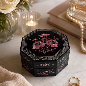 Könnte beinhalten: Eine achteckige schwarze Schmuckschatulle mit einem eingelegten Perlmutt-Blumenmuster in Rosa- und Weißtönen. Der Deckel und die Seiten sind mit komplizierten Blumenmotiven und geometrischen Randmustern verziert und haben eine glänzende Oberfläche.