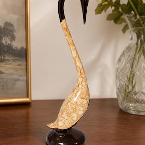 May include: A swan figurine featuring a glossy dark brown head and neck that transitions into a textured, cream and tan crackle-patterned body. The sculpture stands on a smooth, circular dark brown base, showcasing a sleek, elongated S-curve silhouette.