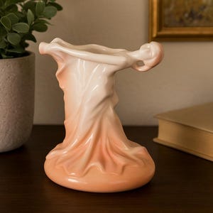 Könnte beinhalten: Eine Keramikvase in fließender, drapierter Form, mit einem Farbverlauf von Pfirsich bis Creme. Die Vase hat ein einzigartiges, künstlerisches Design, das einer Figur in Bewegung ähnelt. Die Gesamtästhetik ist elegant und dekorativ.